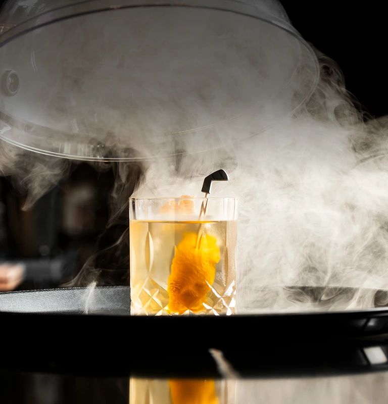 Smoked cocktail revealed under a glass cloche, wisps of smoke curling around a rocks glass.