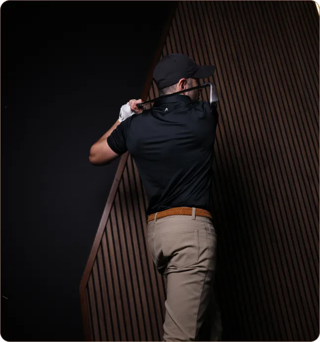 Man in black shirt and cap with beige pants swinging a golf club against a dark background with patterned wooden panels.
