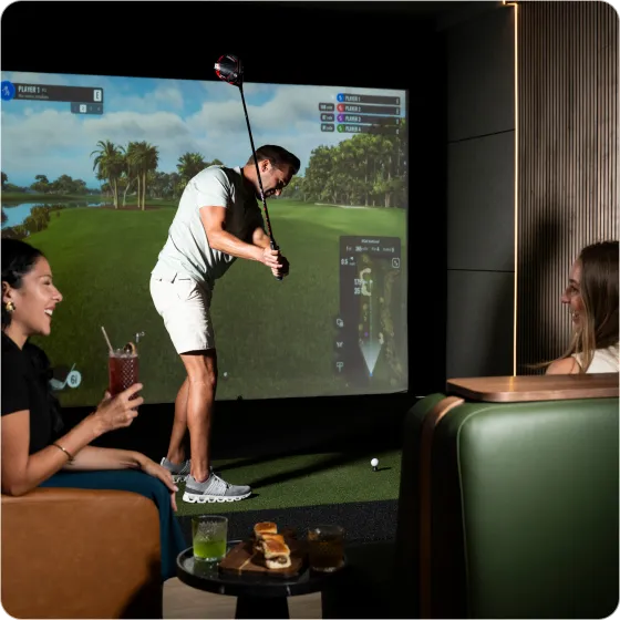 Man swinging golf club in indoor golf simulator while two women watch and enjoy drinks in lounge seating.