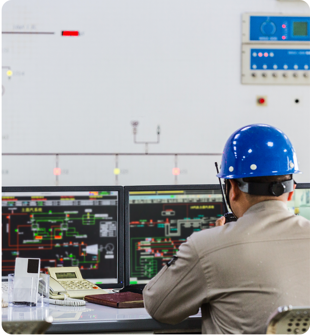 Engineer in a blue hard hat communicates via walkie-talkie while monitoring control system diagrams on dual computer screens.