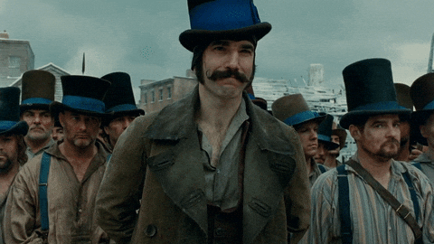 Movie gif. Daniel Day Lewis as Bill the Butcher in Gangs of New York stands menacingly in front of his top hat wearing street gang while they help remove his coat before battle.