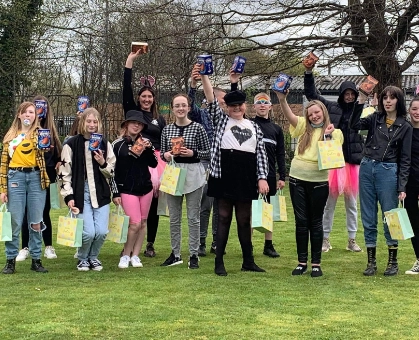 A group of young people posing outdoors on grass, holding up Easter eggs and gift bags, some wearing bunny ears and colourful tutus, at an Easter event.
