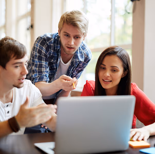 Three young adults gathered around a laptop, discussing and pointing at the screen in a well-lit room.