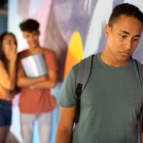 Sad young man with a backpack looking down while two blurred teenagers talk in the background.
