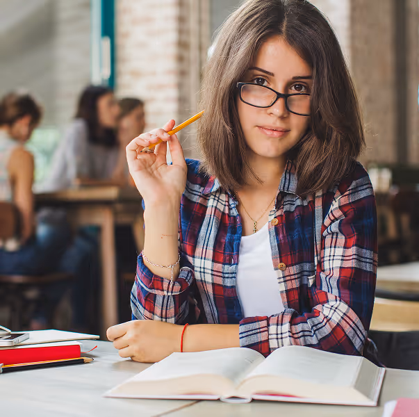 Young woman wearing glasses and a plaid shirt holding a pencil while sitting at a table with an open book in a library or study space.