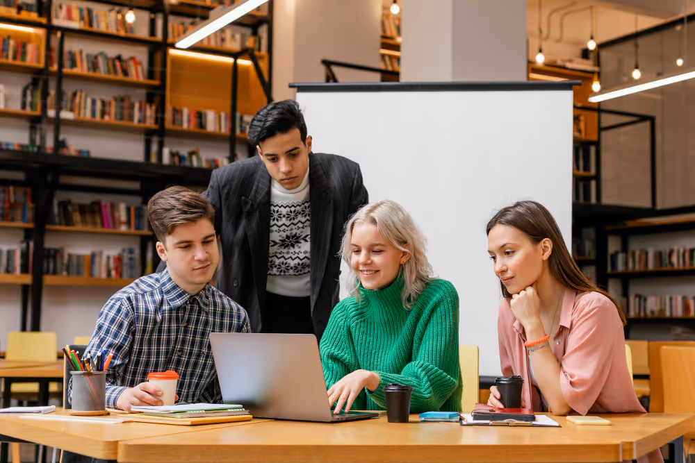 Four young adults sitting and standing around a laptop in a modern library, discussing and smiling.
