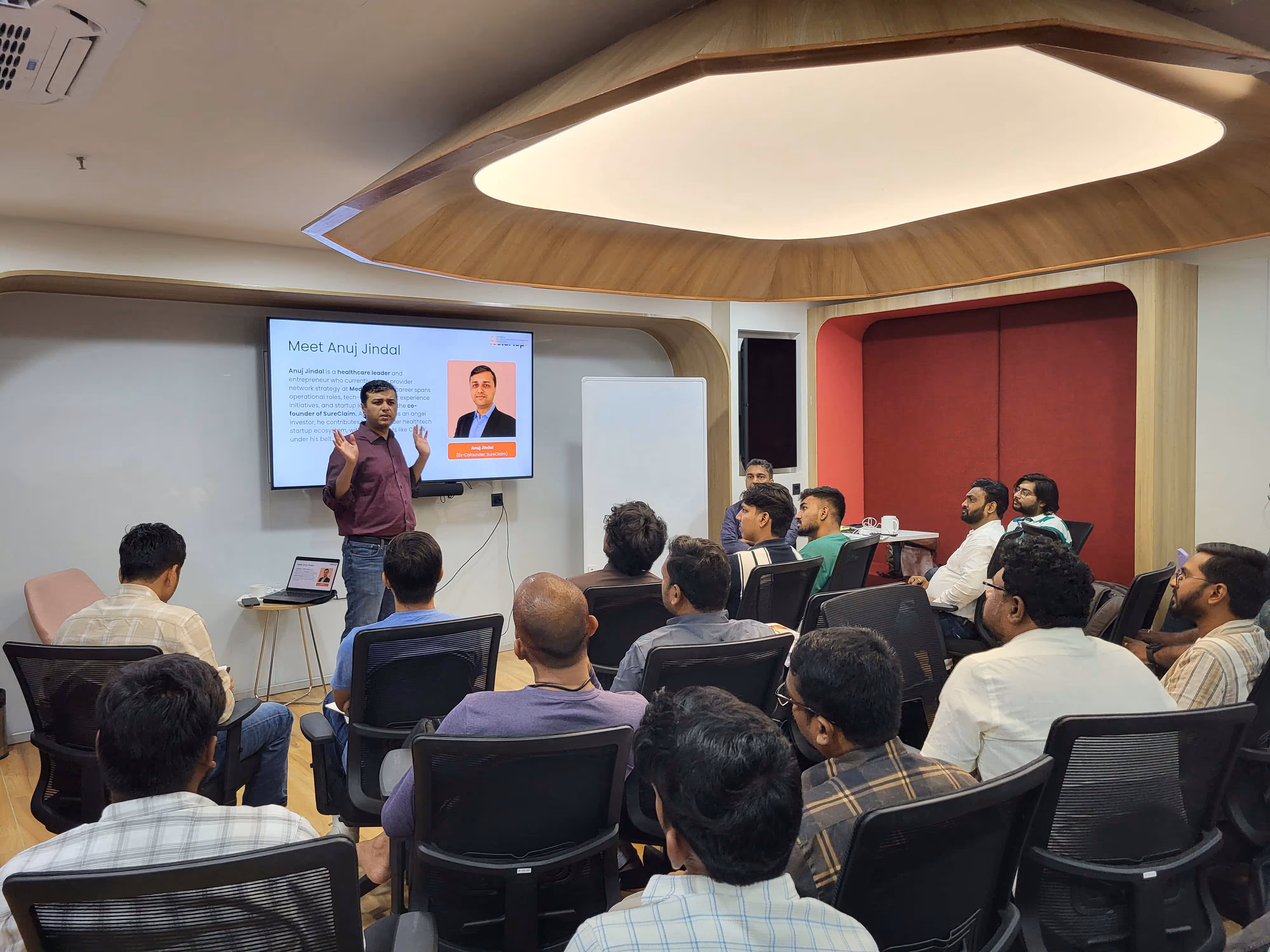 Young man wearing headphones and a pink sweatshirt writing with a pencil in a notebook in a library setting, with another person working on a laptop in the background.