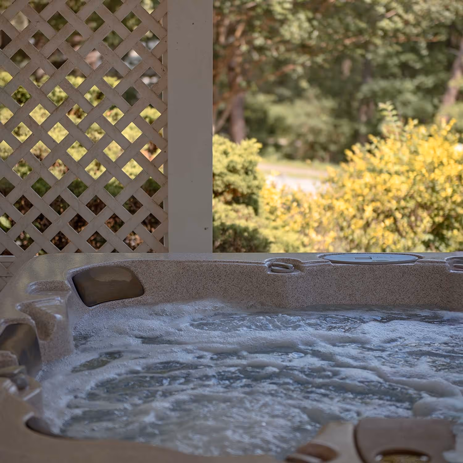Outdoor hot tub with bubbling water, partially enclosed by a white lattice privacy screen, surrounded by lush greenery and sunlit bushes in the background.