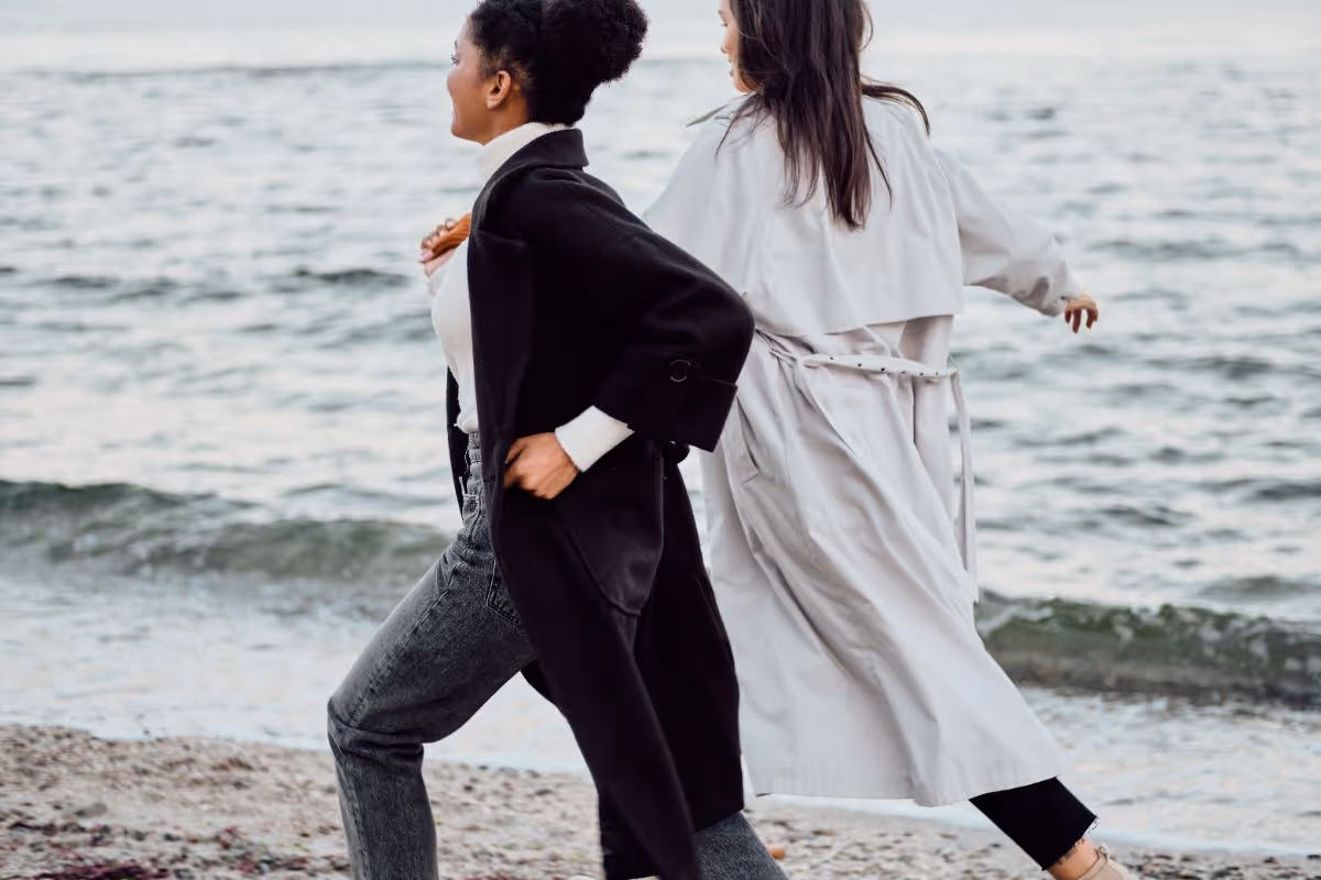 Two women walking briskly along a beach near the water, one wearing a black coat and the other a light trench coat.