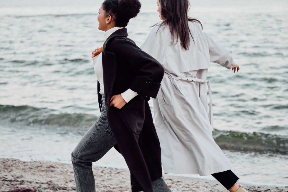 Two women walking briskly along a beach near the water, one wearing a black coat and the other a light trench coat.