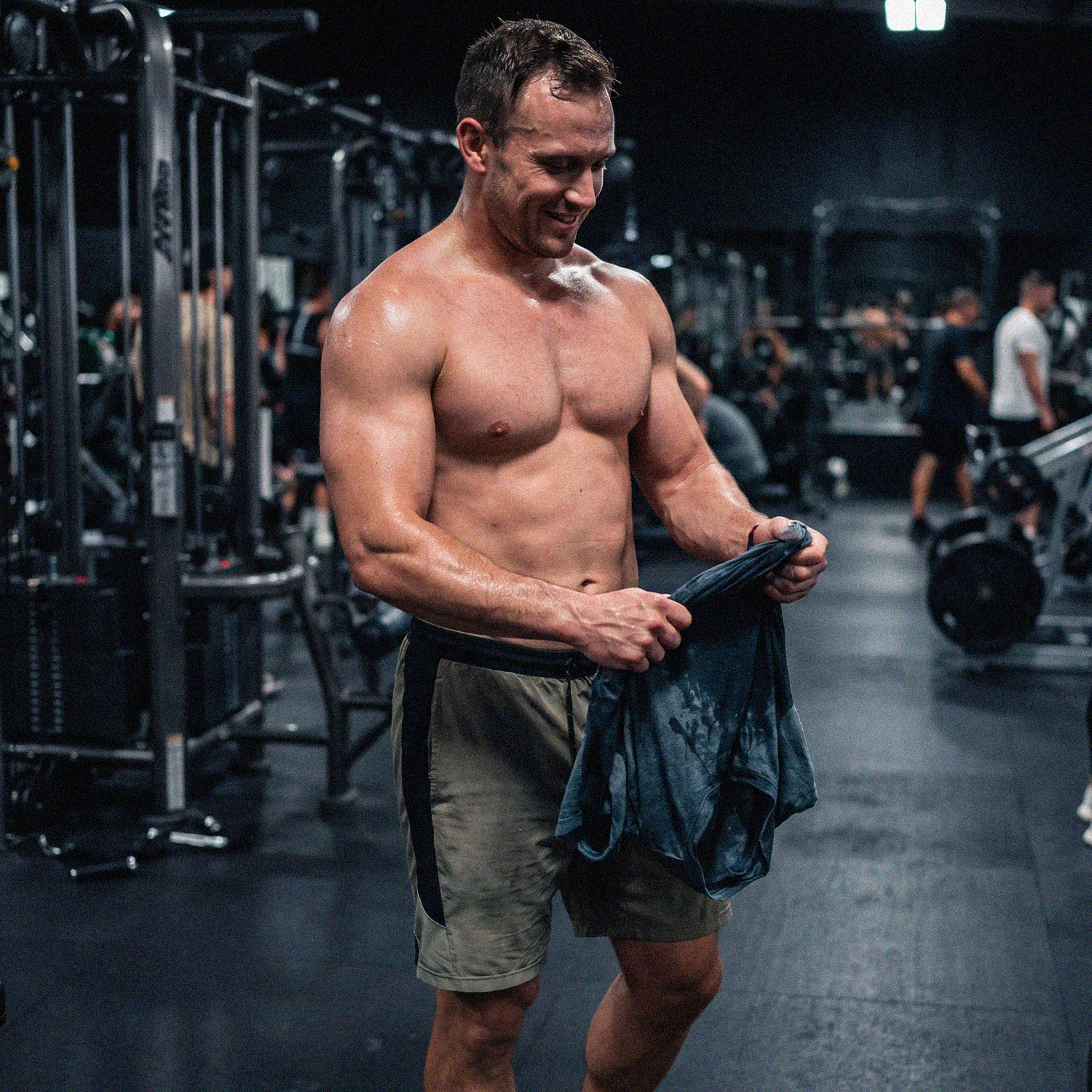 Muscular shirtless man holding a wet shirt in a busy gym with workout equipment in background.