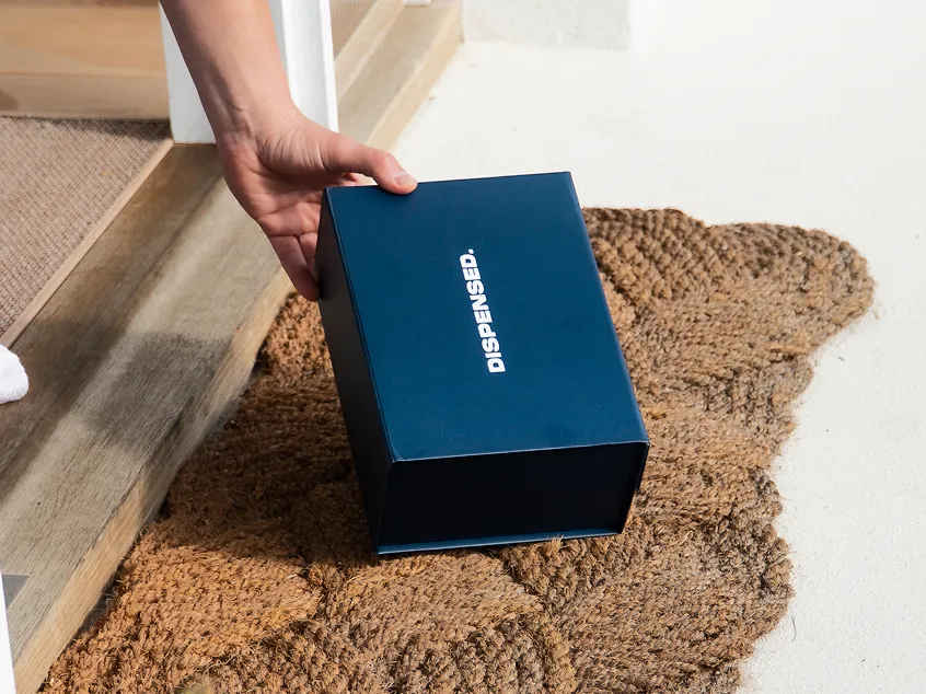 Close-up of a Dispensed delivery box being placed on a front doorstep, sitting on a door mat and being picked up by a patient.