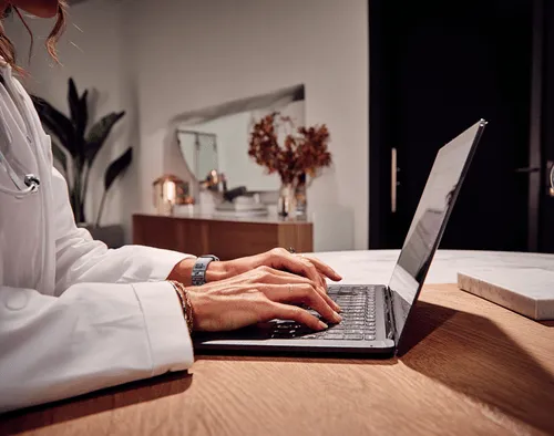 A clinician in a lab coat is sitting at their desk typing on their computer in a well lit home office environment.