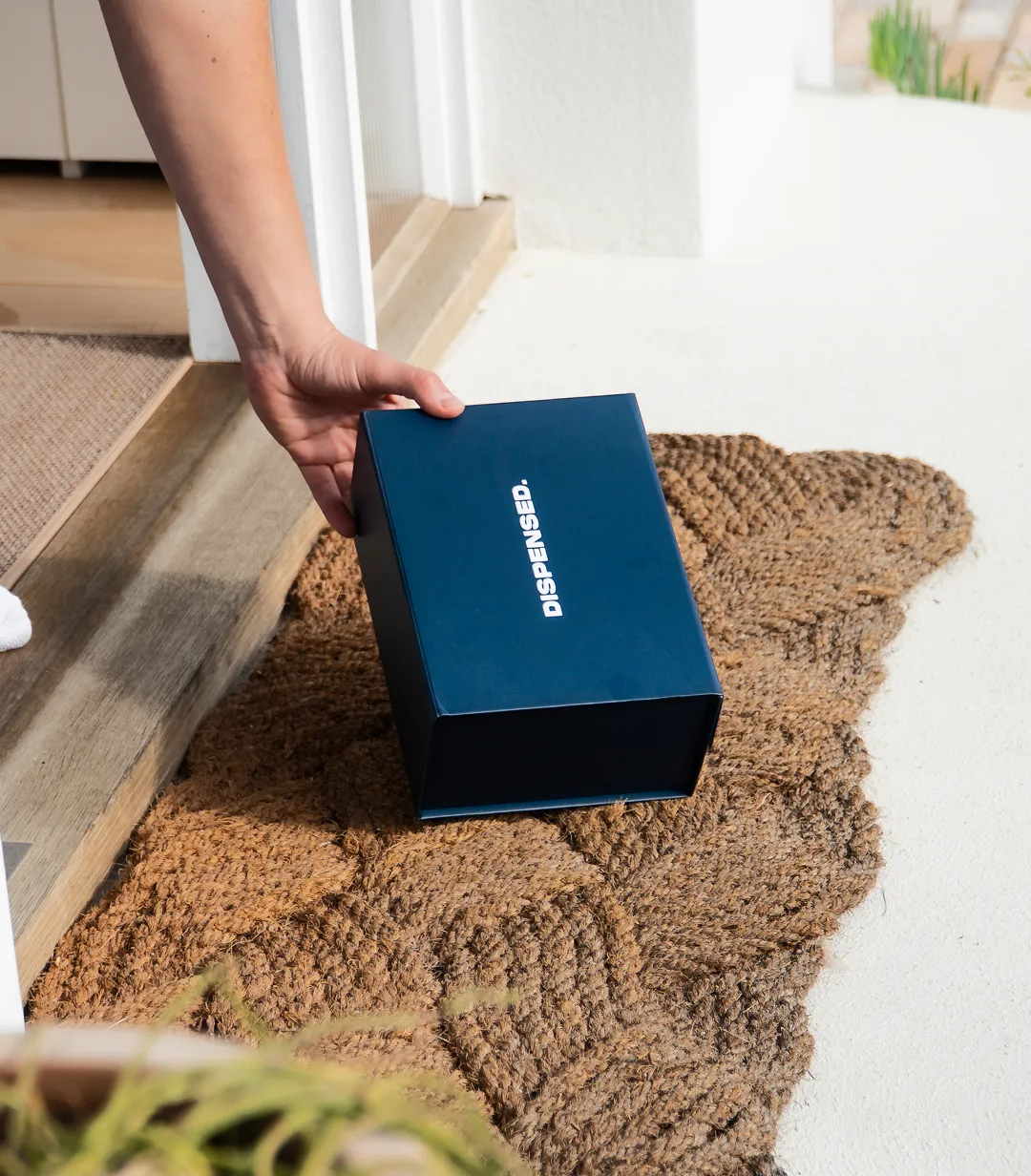 Close-up of a Dispensed delivery box being placed on a front doorstep, sitting on a door mat and being picked up by a patient.