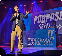 Man speaking on stage with microphone in hand, standing next to a sign that reads 'Purpose Con Ty Thompson' under purple and red stage lights.