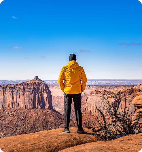 Person in yellow jacket standing on rocky cliff overlooking a vast desert canyon landscape under a clear blue sky.