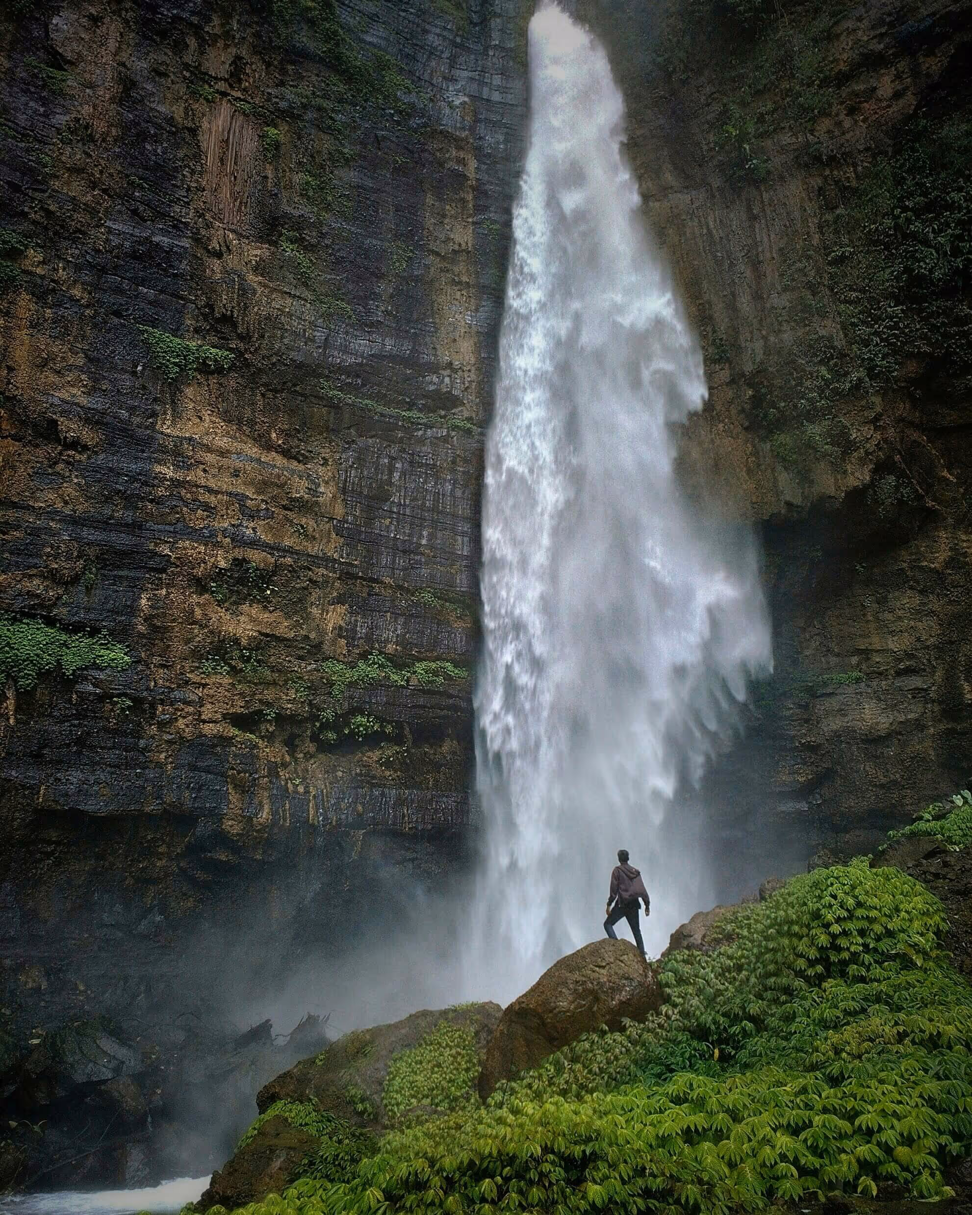 Person standing on a rock near a tall, powerful waterfall surrounded by steep cliffs and green vegetation.
