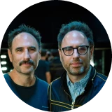 Two men standing close together, one with a mustache and short hair, the other wearing glasses and a patterned scarf.