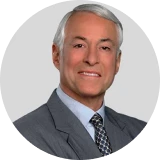 Portrait of a smiling middle-aged man with gray hair wearing a gray suit and patterned tie against a plain gray background.