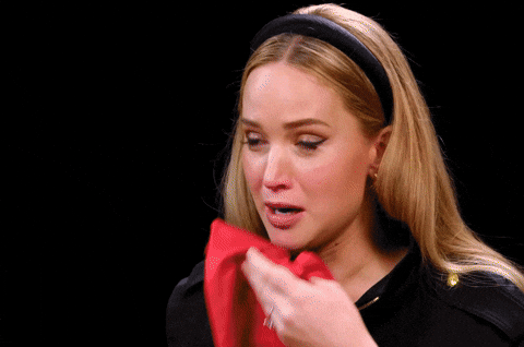 TV gif. Jennifer Lawrence sits for an interview on the talk show "Hot Ones." A delirious Lawrence has tears in her eyes after eating a particularly spicy hot wing. She wipes her mouth with a red napkin as she repeatedly asks "What do you mean?" while laughing, then crying.