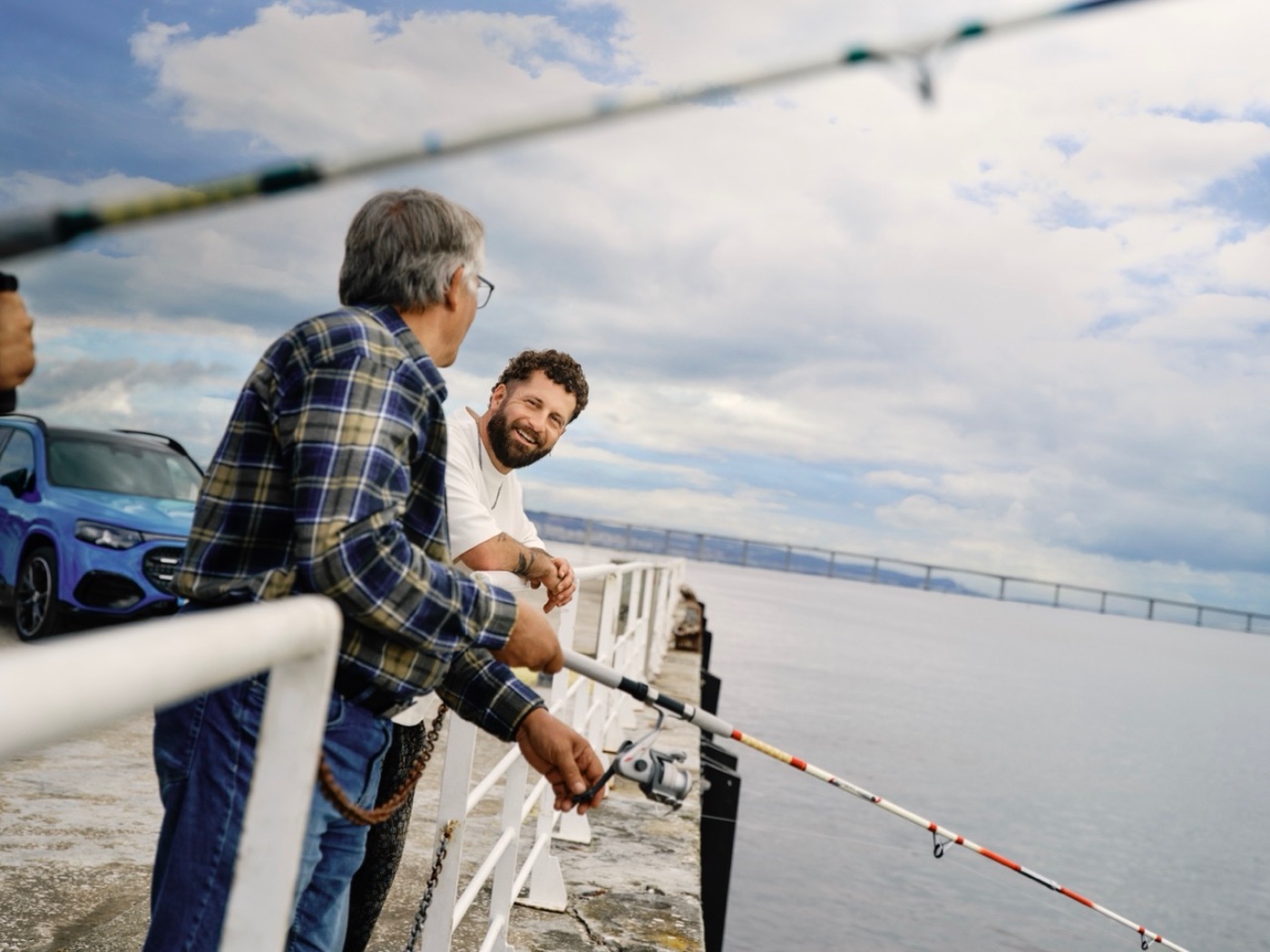 Two men fish over a pier with a blue electric vehicle in the background