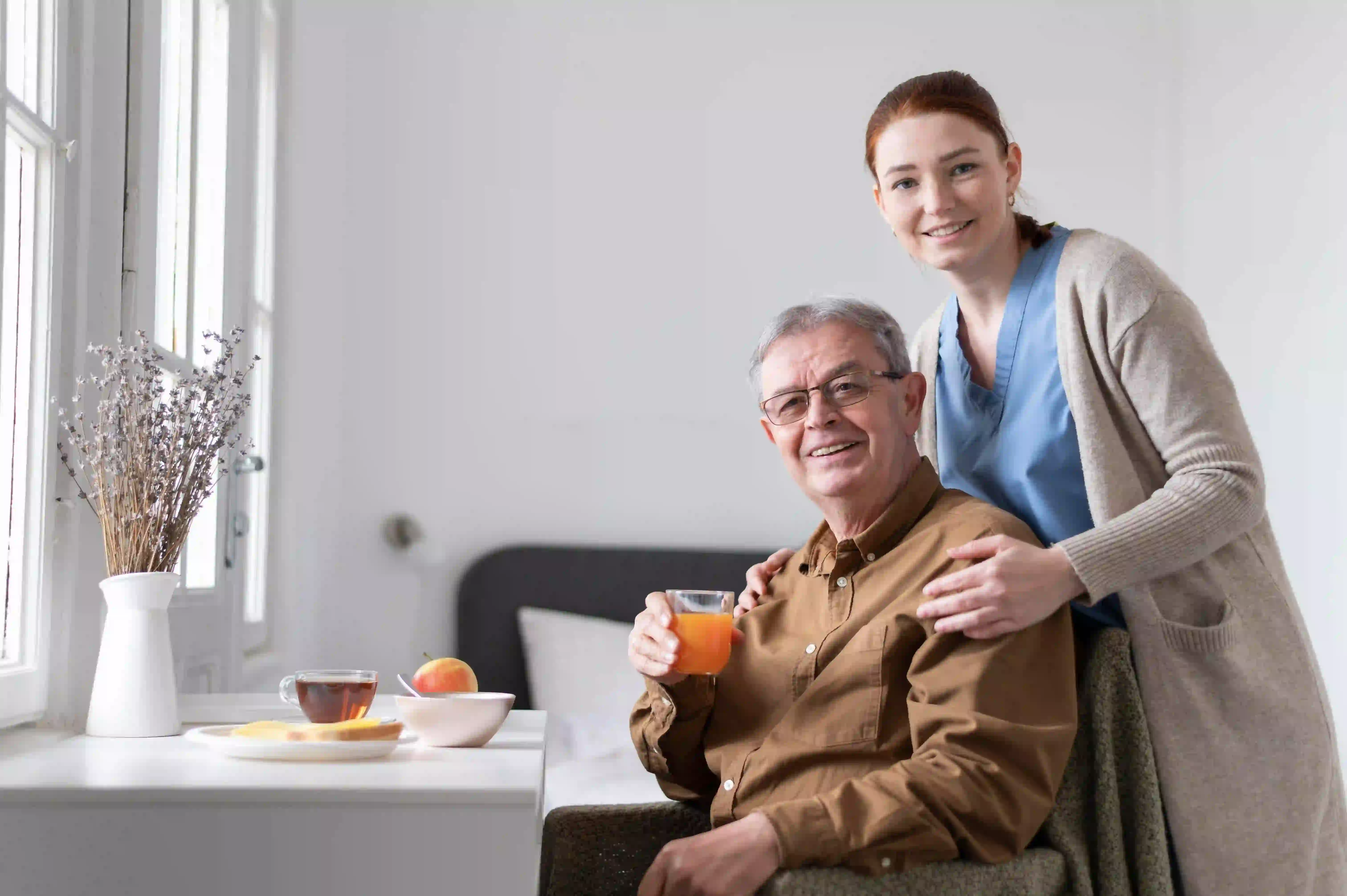 Caregiver standing beside senior man enjoying breakfast at home.