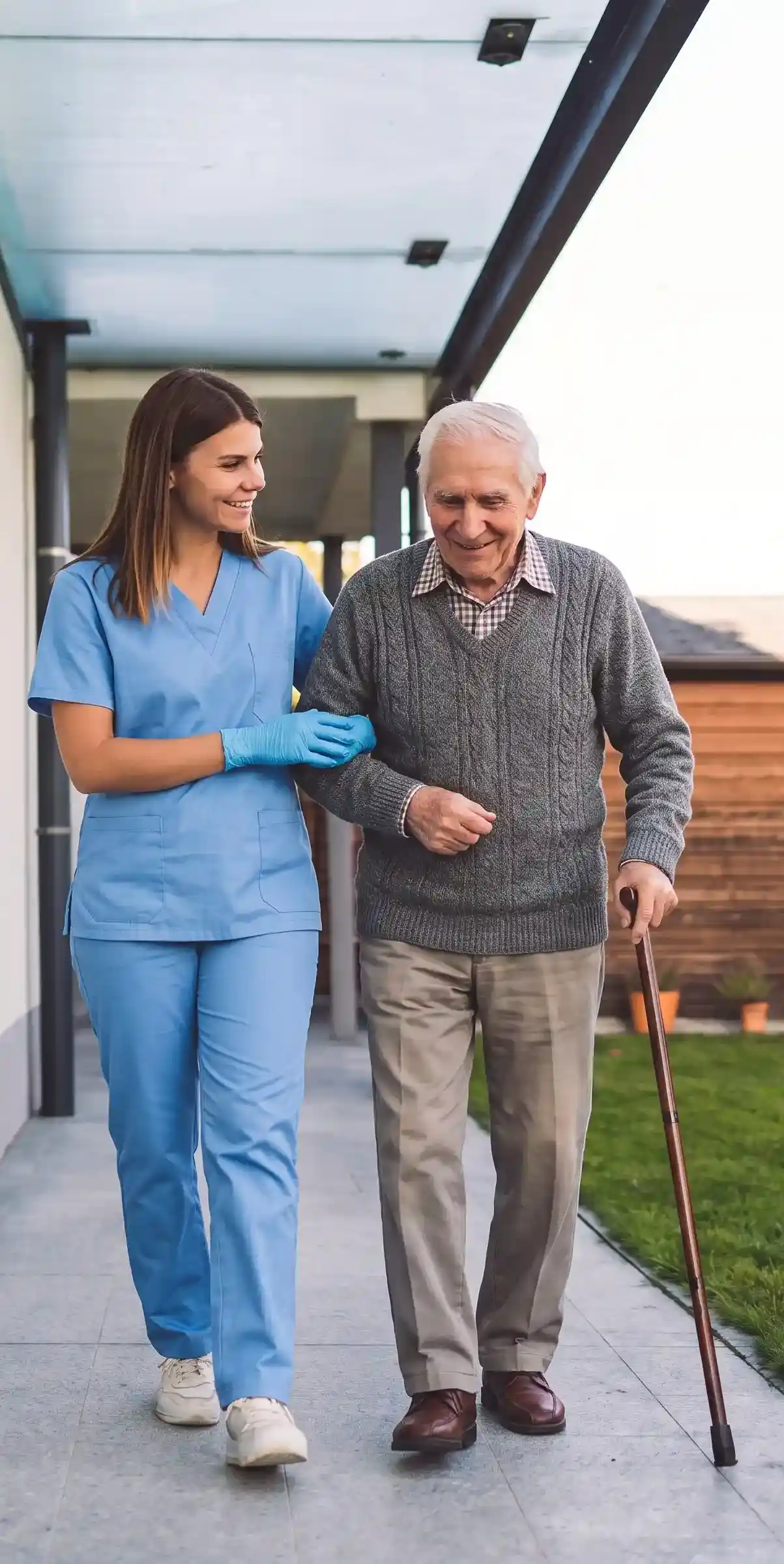 A female caregiver in blue scrubs assisting an elderly man with a cane walking safely outside.