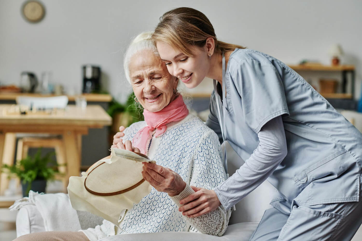 Smiling senior woman embroidering and showing her handicraft to caregiver
