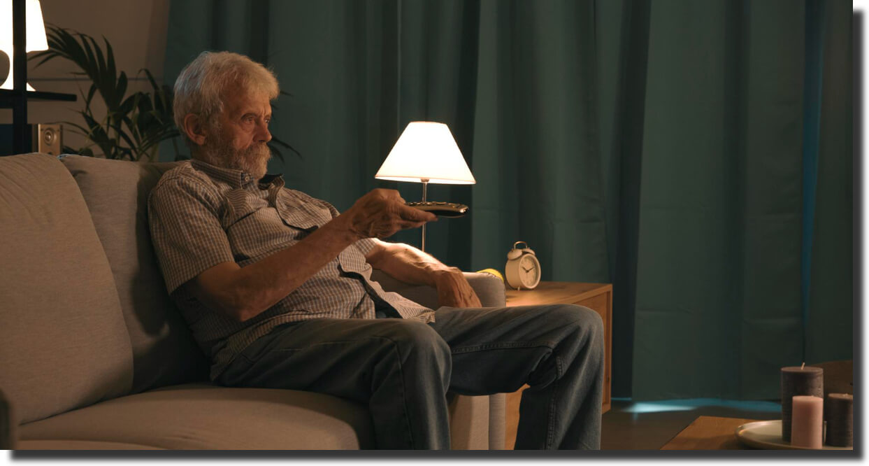 Older man on sofa turning on a lamp at night to improve lighting for safer dementia care environment.