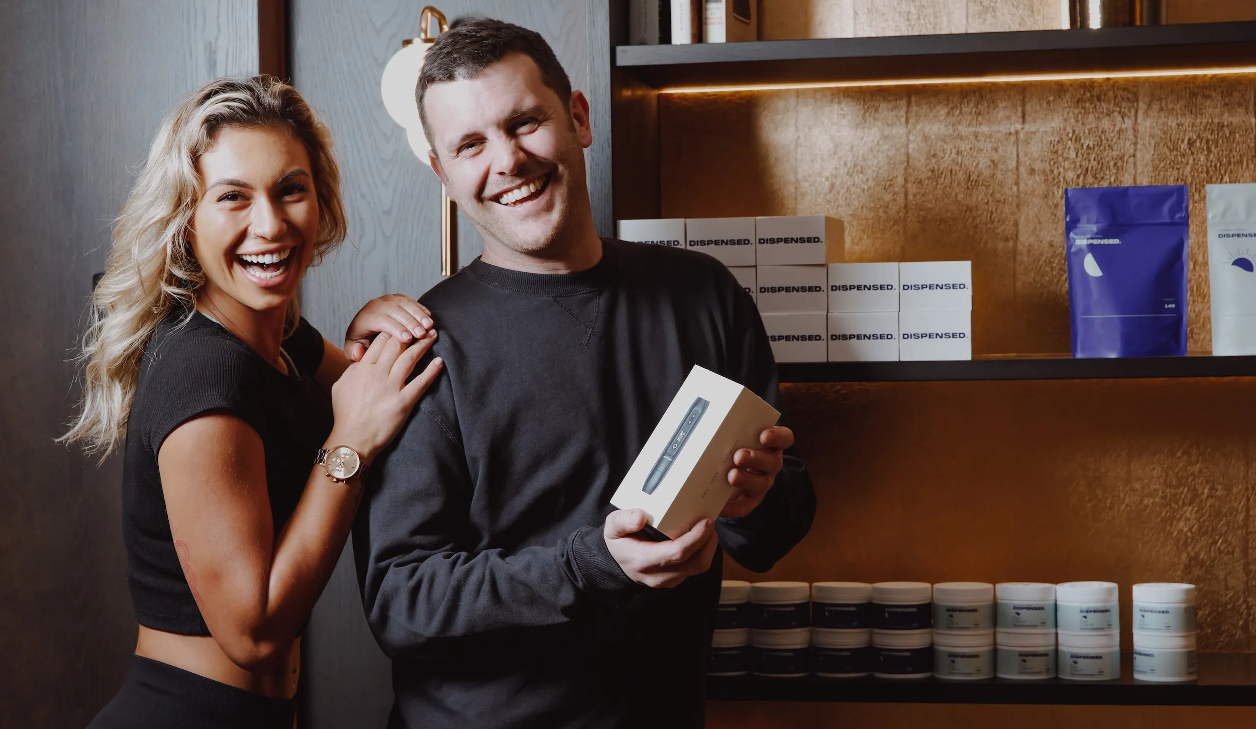 Woman smiling with her hands resting on the man’s shoulders as he holds a Dispensed-branded package, standing together in front of shelves stacked with product boxes.