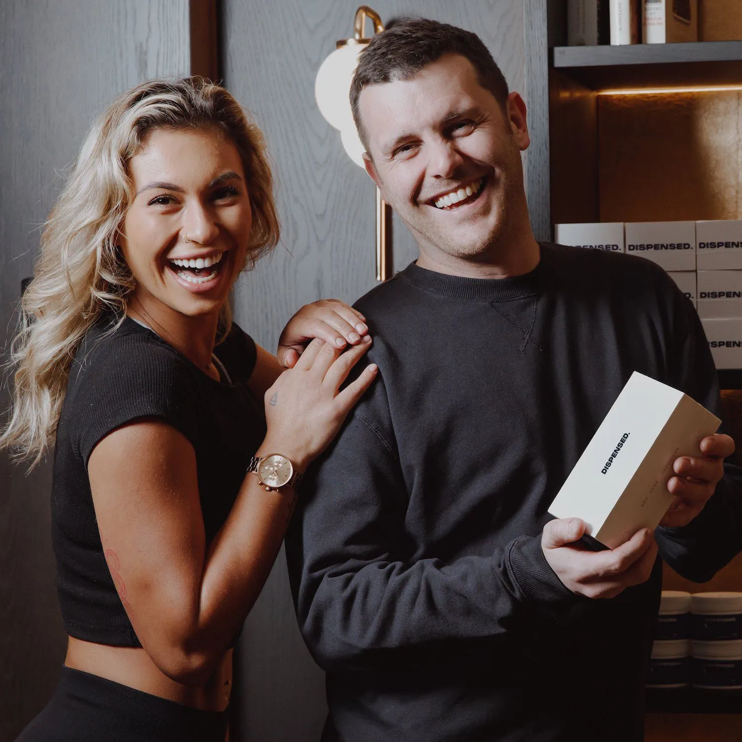 Man and woman in front of a shelf stocked with Dispensed products, the man is holding a medical box and the woman is smiling.