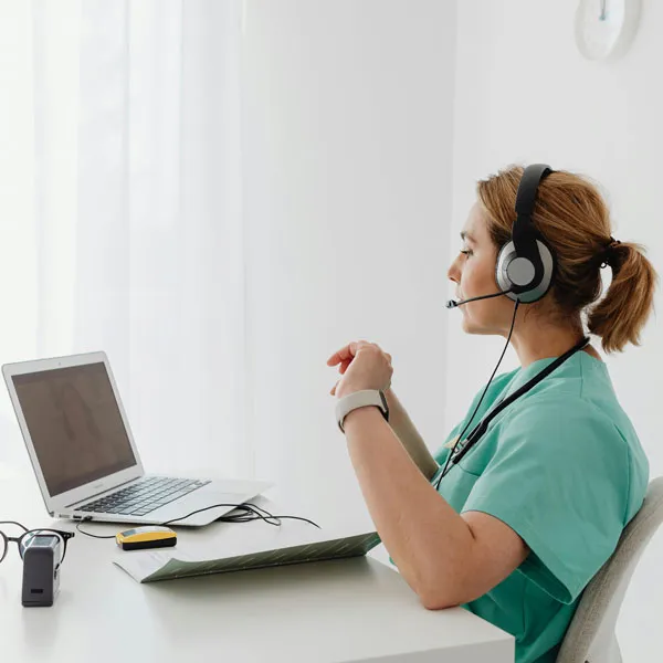 A woman in scrubs who appears to be a nurse with a headset on conducting a telehealth consultation on her computer at a desk in a well lit office.