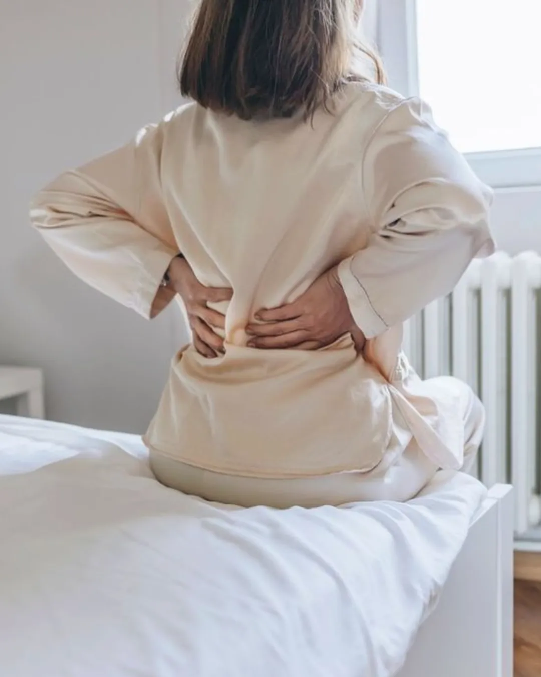 A woman in pyjamas getting out of bed whilst holding her back which appears to be in pain.