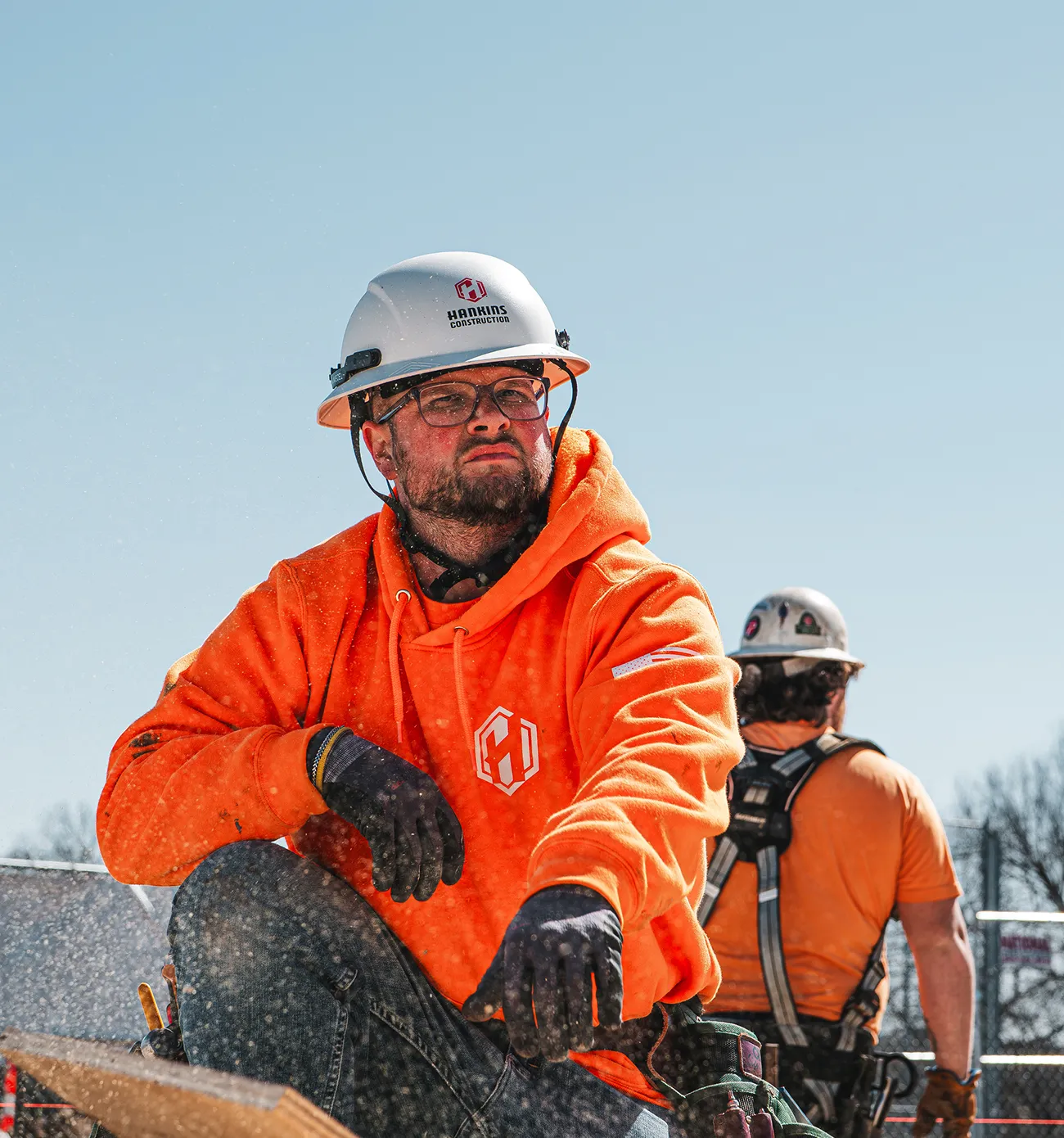 Construction worker in orange hoodie and white hard hat sitting on a wooden plank at a construction site with another worker in an orange shirt and harness in the background.