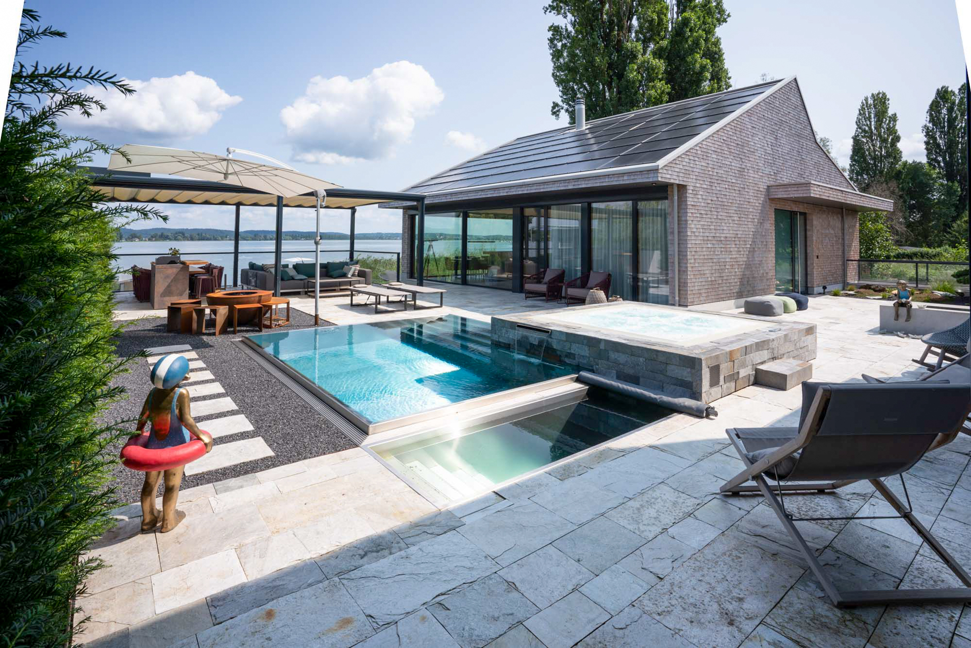 Haus am See mit Naturstein, Infinitypool und Pergola mit Sitzgruppe und Blick auf den See. 