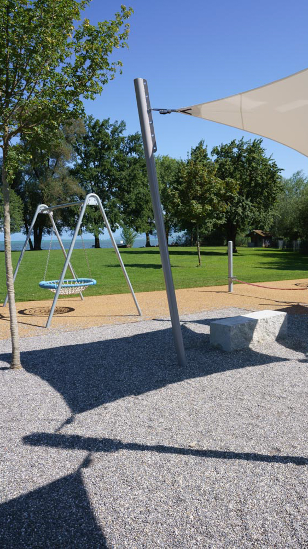 Spielplatz mit Nestschaukel, Sonnensegeln und Sitzquader unter freiem Himmel – umgeben von Rasenfläche, Bäumen und Blick auf den Bodensee im Hintergrund.“