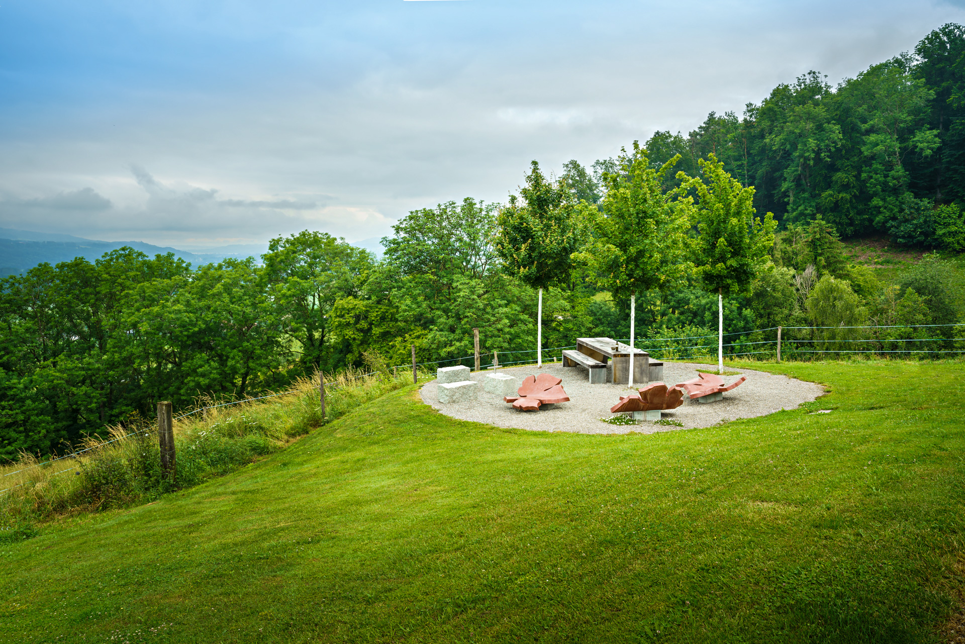 Rastplatz mit Kunstwerk Sitzgelegenheiten und Tisch / Bank aus Stein inmitten einer Grünen wiese und vielen Bäumen im Hintergrund. 