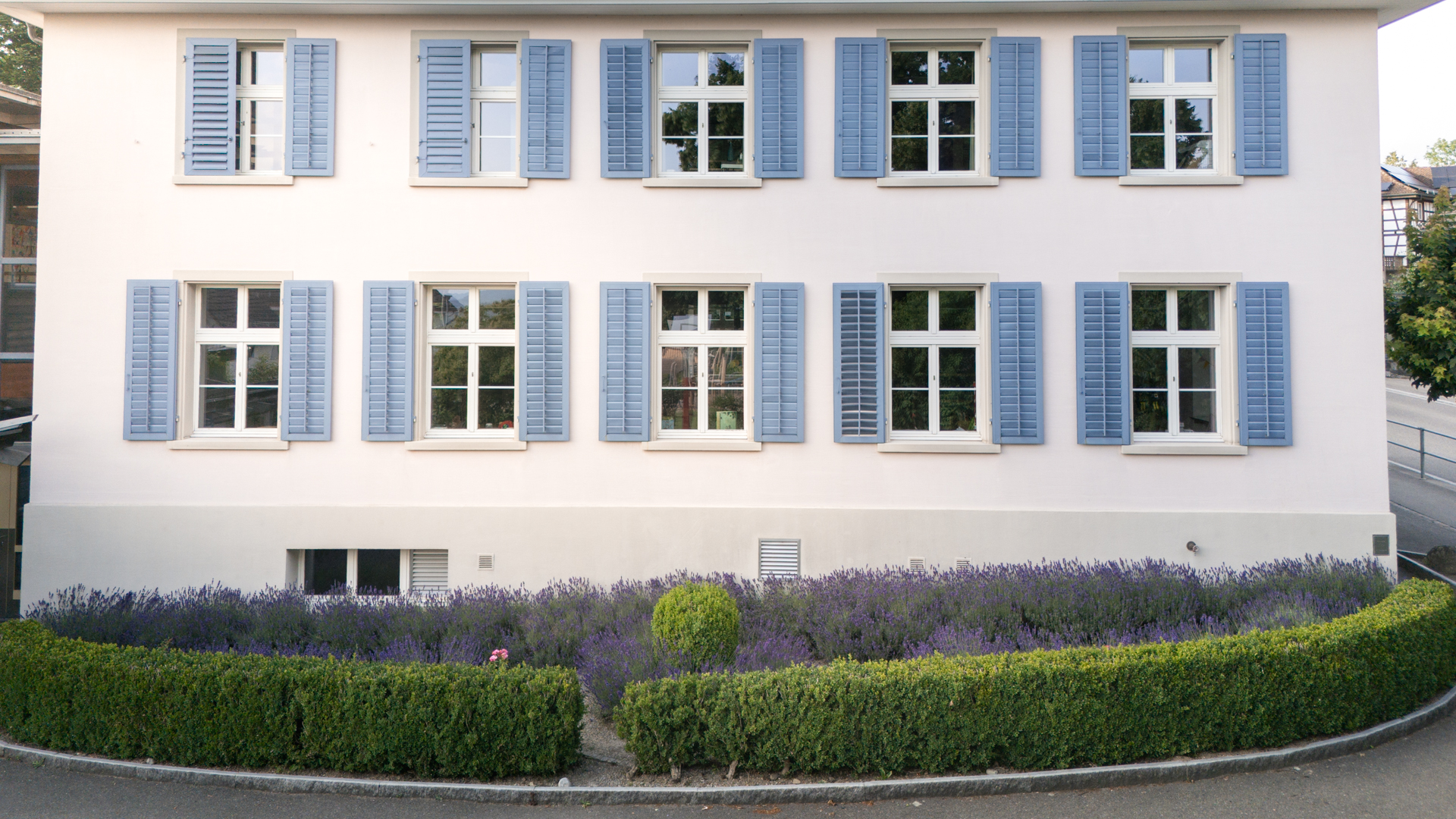 Kindergarten: grosses Haus mit weisser Fassade und hellblauen Fensterläden, davor ein Blumenbeet mit blühendem Lavendel und einer kleinen grünen Hecke. 