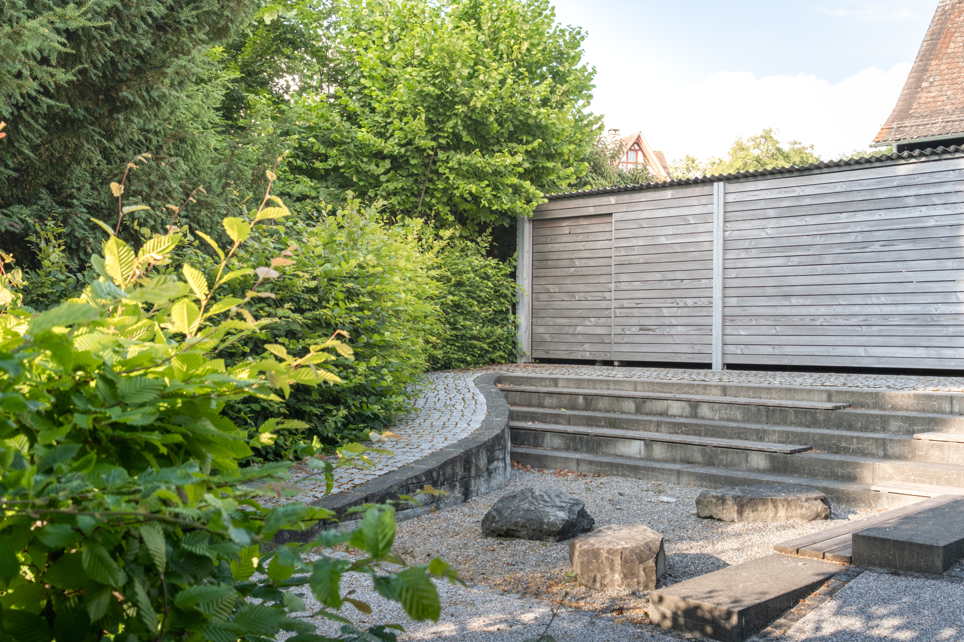Kiesplatz mit Bettontreppe, Stein und Holz Sitzgelegenheiten umgeben von grünen Hecken. 