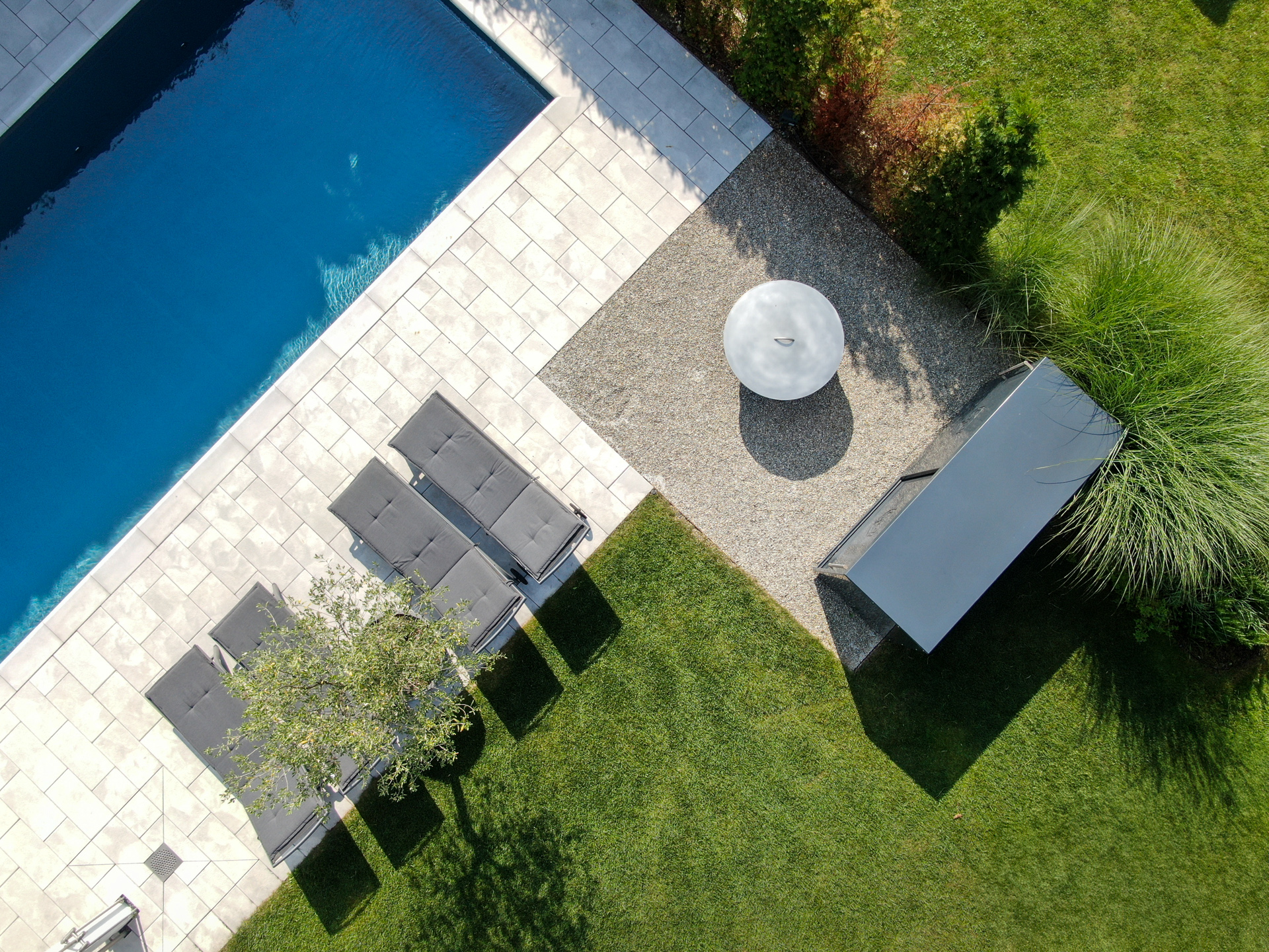 Drohnenaufnahme von Swimmingpool mit Naturstein und vier Liegestühlen, daneben Rasen mit Gräsern. 