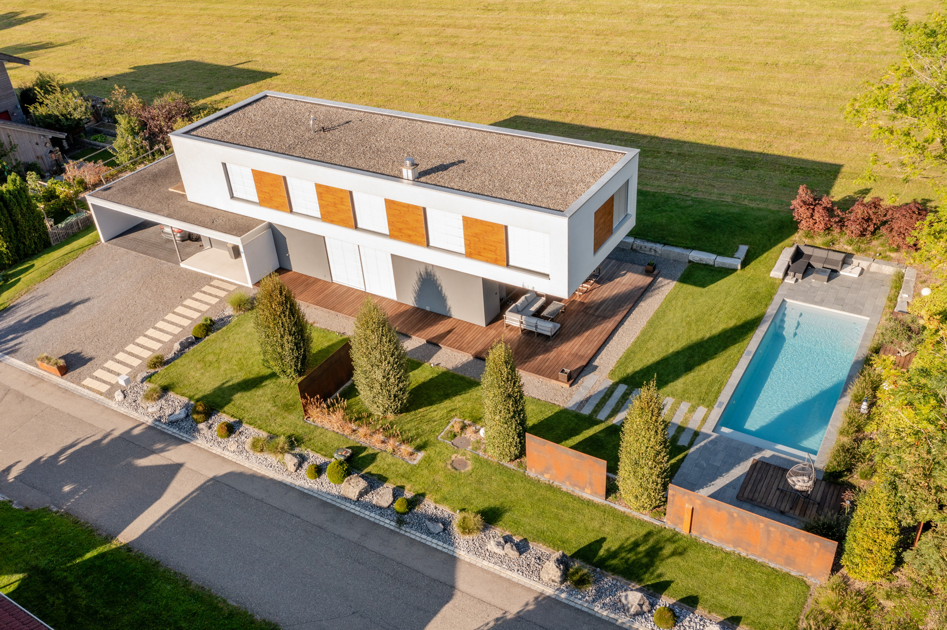 Drohnenfoto eines modernen Hauses am Feldrand, mit grossen Bäumen und einem Infinity Swimmingpool. 