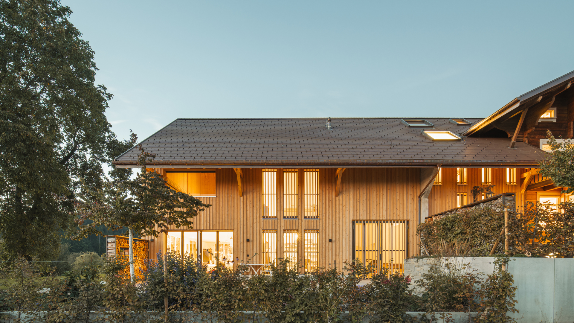 Beleuchtetes Wohnhaus mi Holzverschalung und Betonhochbeetne und einem grossen alten Baum. 