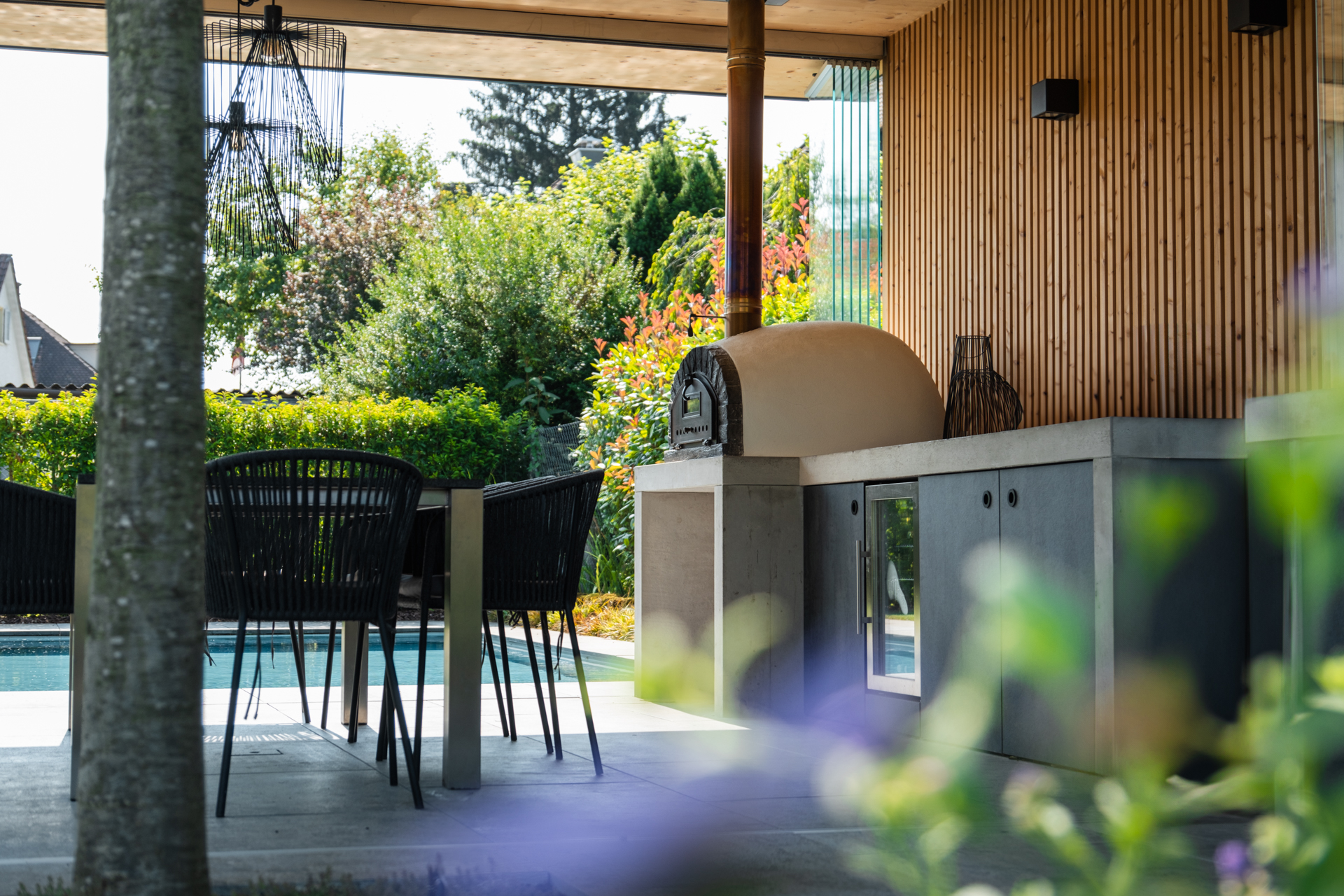 Tiefenunscharfes Bild im Vordergrund violette Pflanzen unscharf im Hintergrund eine Loggia mit Pizzaofen, weiter hinten nicht gut sichtbar ein Swimmingpool. 