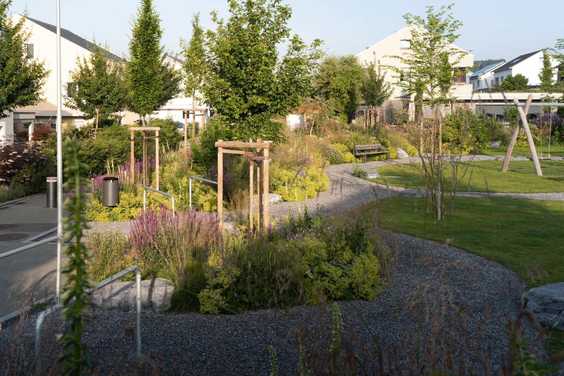 Moderner Quartierpark mit geschwungenen Kieswegen, Blühpflanzen, jungen Bäumen, Sitzbänken und Spielgeräten, eingebettet zwischen Wohnhäusern.