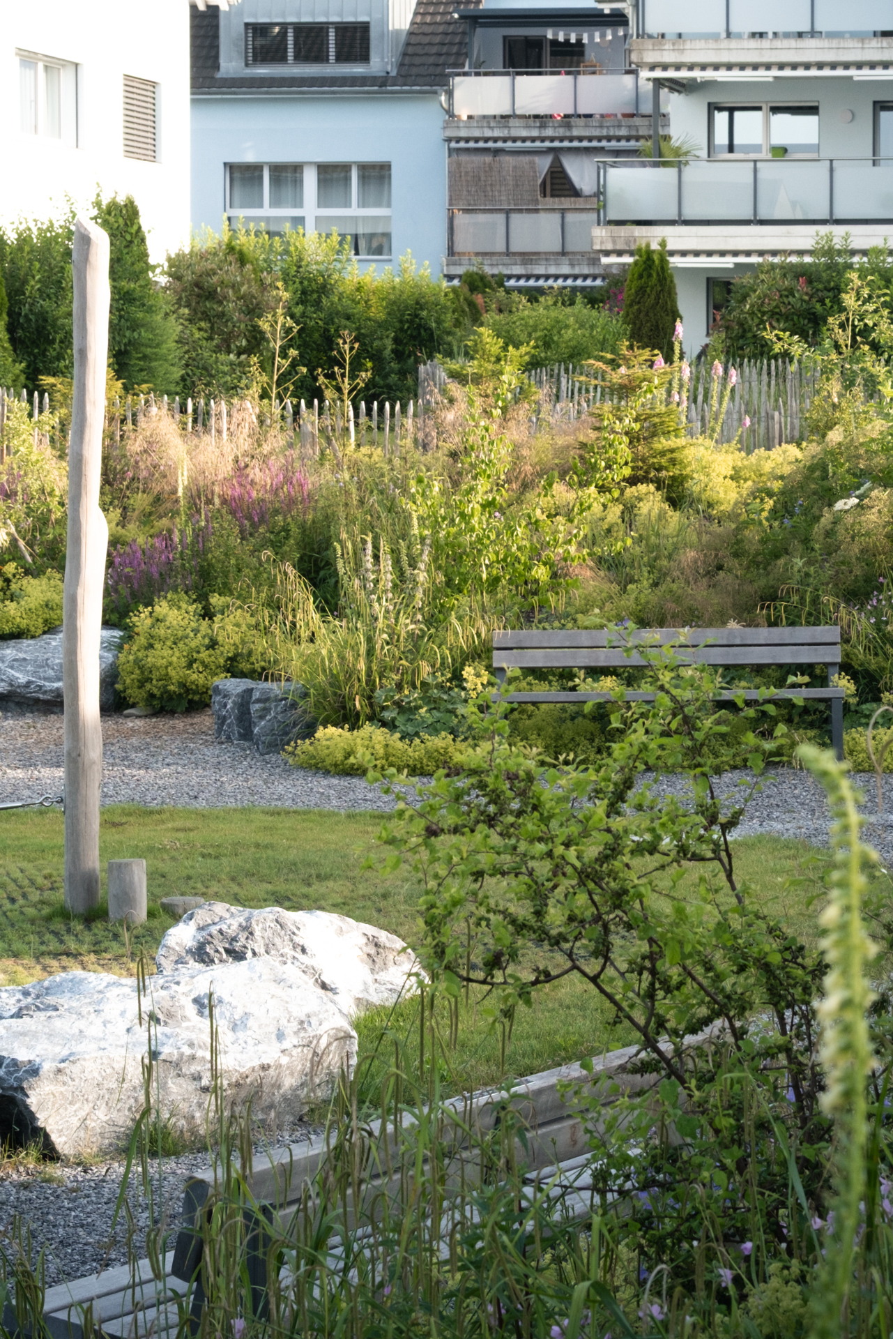 Sitzbank im begrünten Quartierpark mit Blühstauden, Ziersträuchern, Natursteinen und Kiesweg, umgeben von Wohnhäusern.