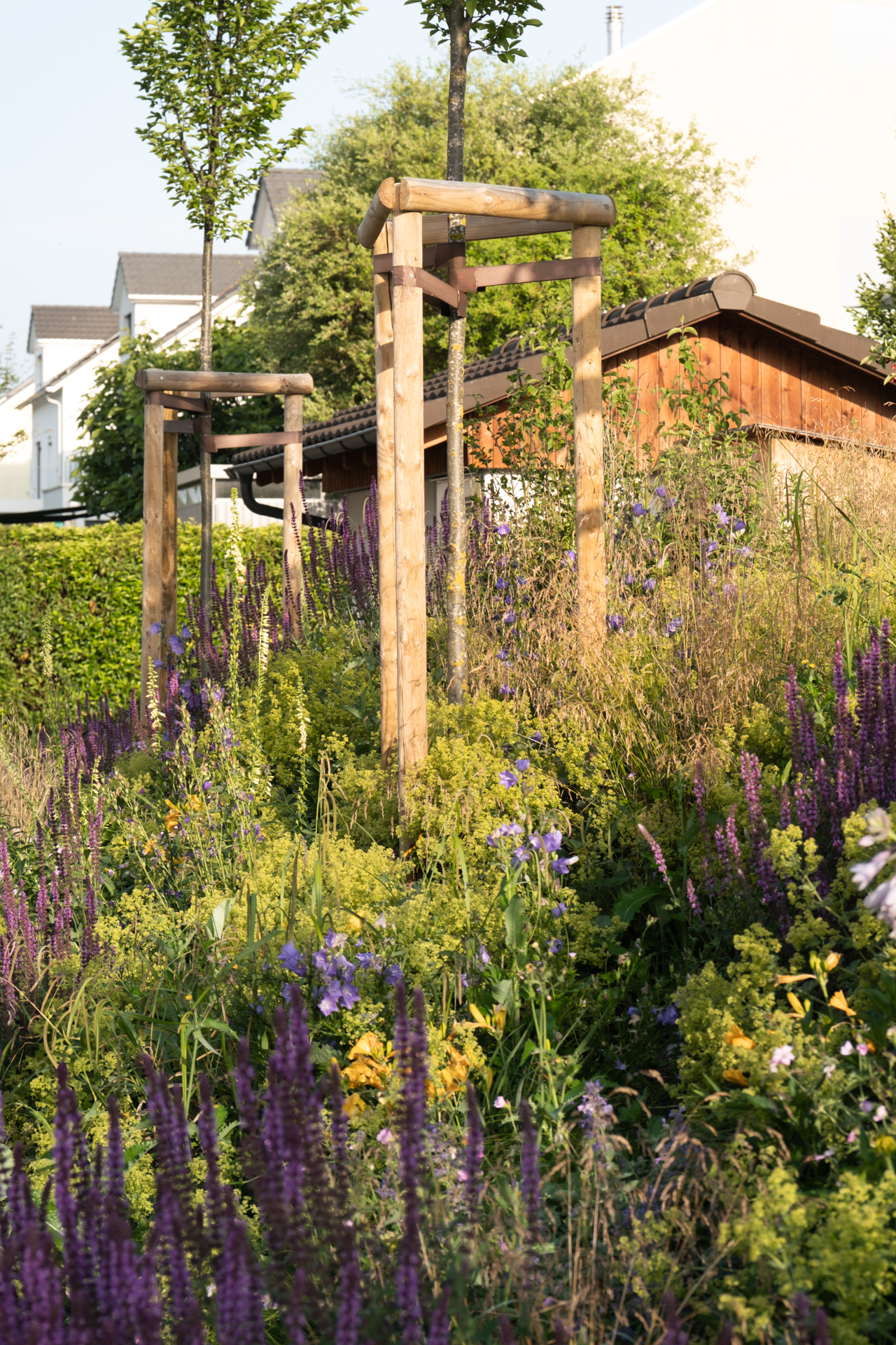 Blühendes Blumenbeet mit hochstämmigen Bäumen im Hintergrund un deiner Holzhütte.