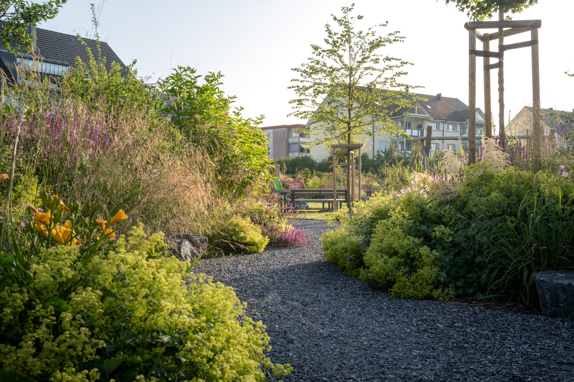 Geschwungener Kiesweg in mitten von üppiger Begrünung im Hintergrund Wohnhäuser.