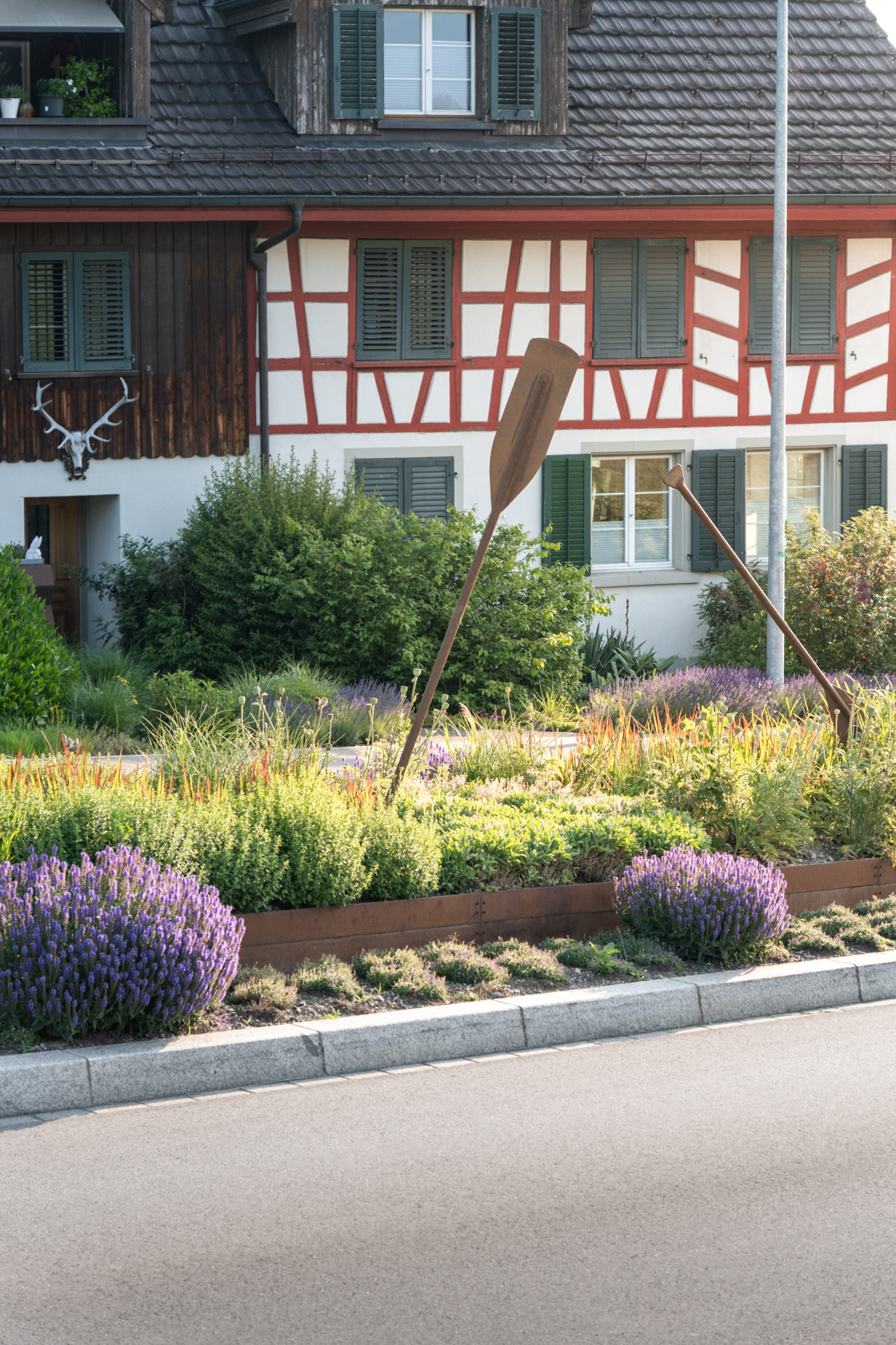 Riegelhaus im Hinterrgudn im Vordergrund eine Verkehrsinsel im nautischen Style mit Stahl Rudern und farbiger Bepflanzung.