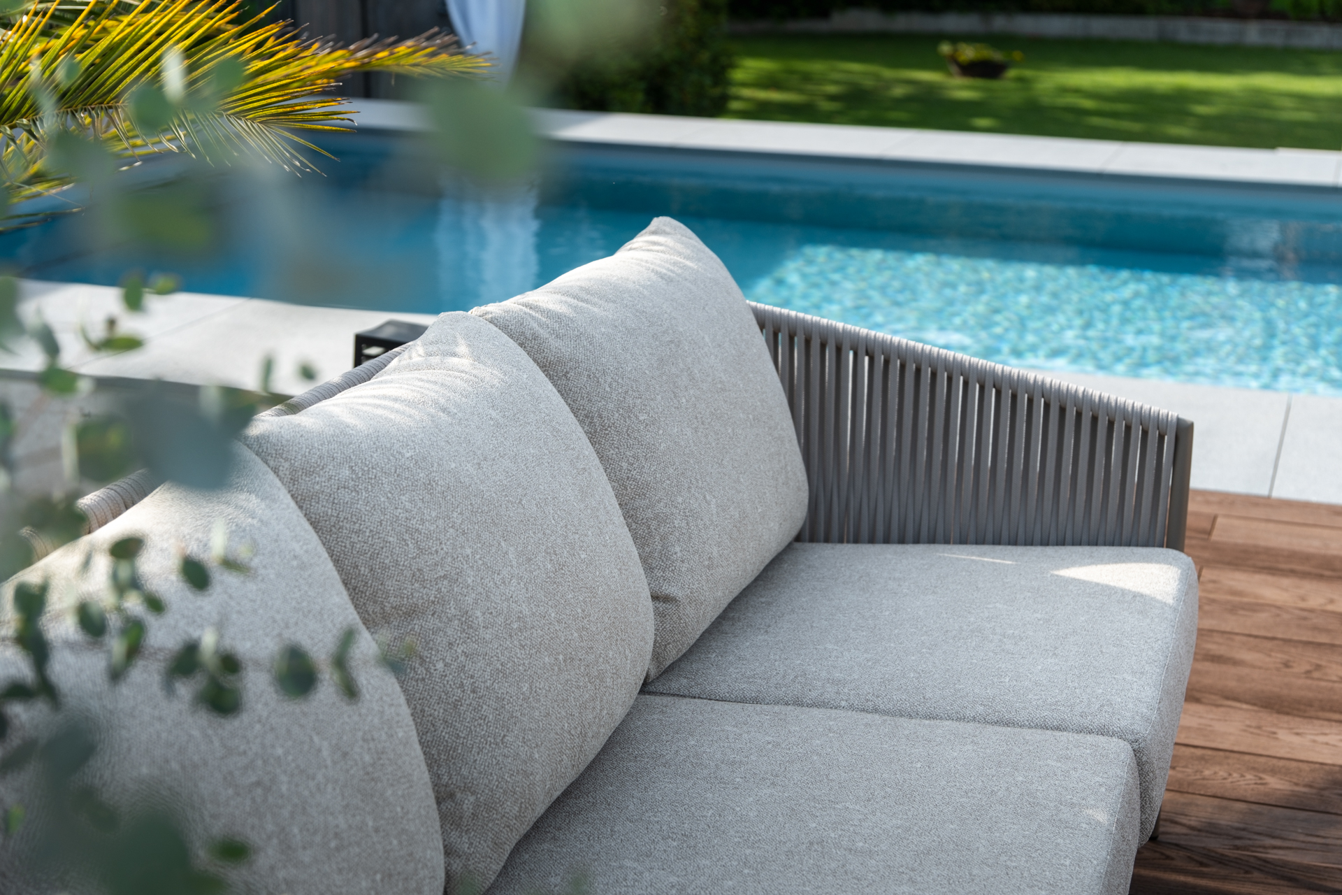 Graues Garten Lounge Sofa mit Palmenwedel und Swimmingpool im Hintergrund. 