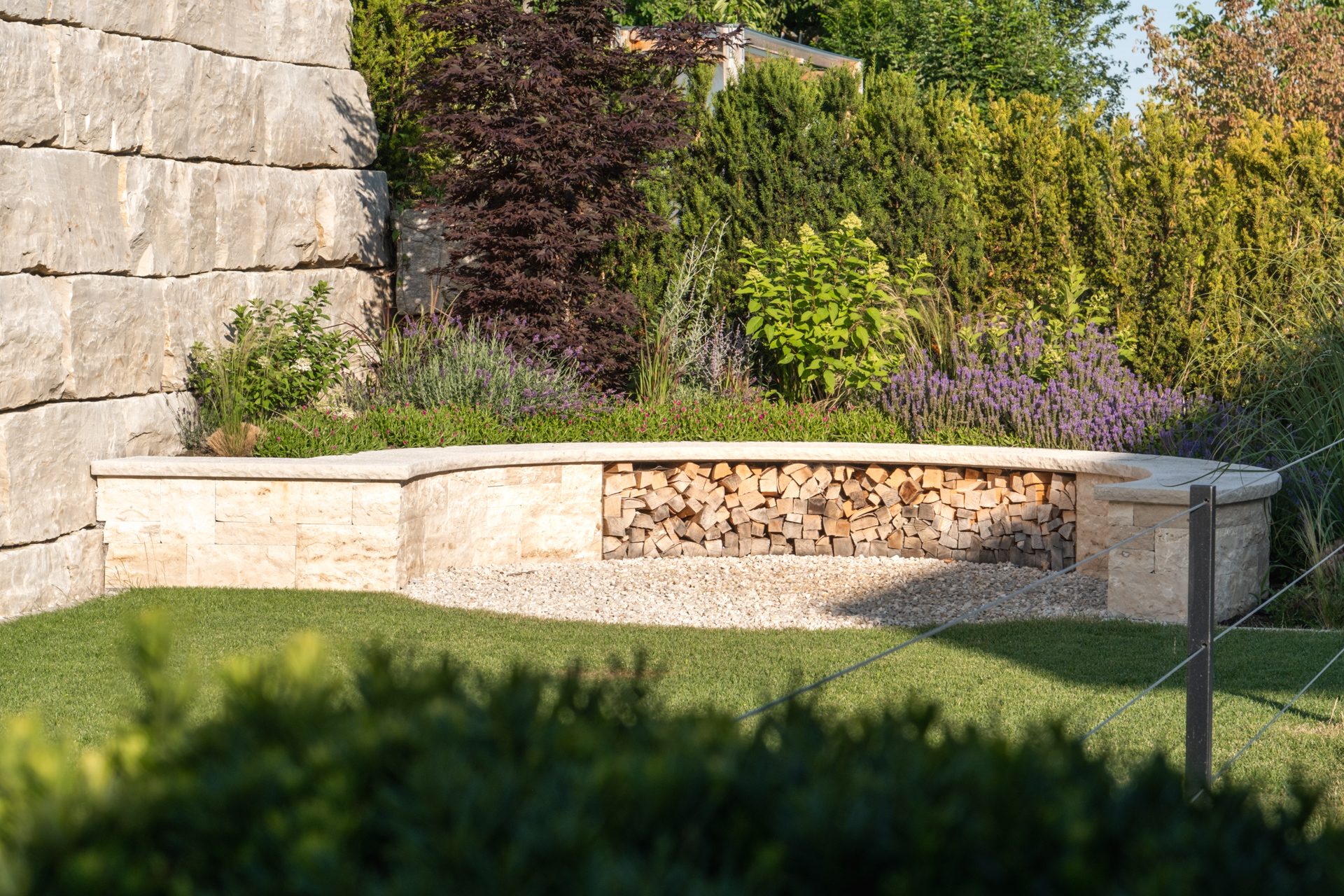 Runde Sitzbank aus Naturstein dient als Holzlager; im Hintergrund eine harmonisch abgestimmte Begrünung. 